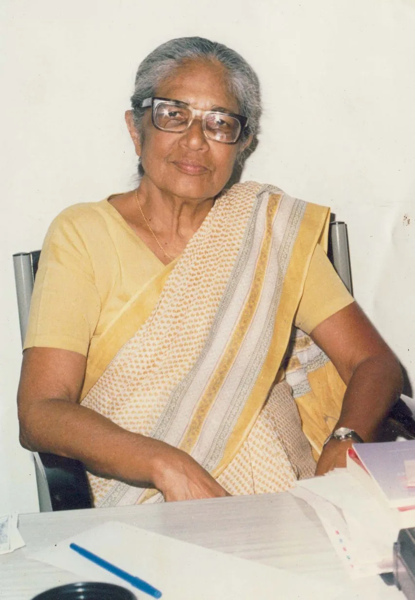 Portrait of Anna Mani in later years, seated at her desk in a yellow sari